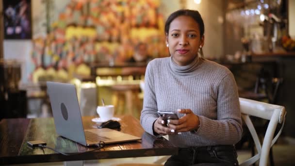 非洲裔美国女商人在咖啡吧餐厅与笔记本电脑一起工作时看着相机。使用计算机的妇女远程工作者坐在桌子上，在室内喝咖啡 — 图库视频影像