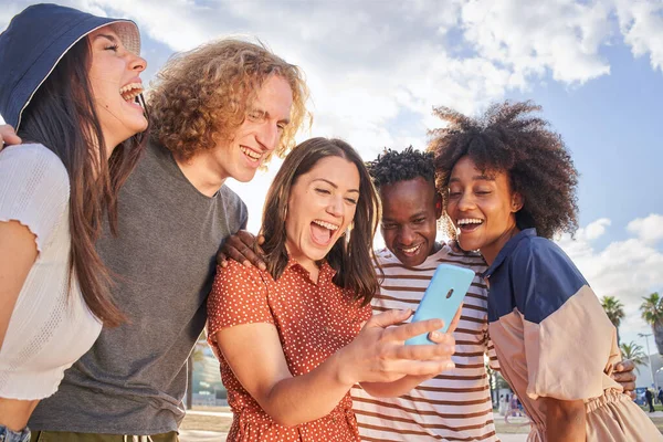 Grupo de jóvenes que se divierten viendo algo en sus teléfonos celulares. Los chicos se abrazan y se ríen entre sí. Concepto de multiétnica, amistad, tecnología. — Foto de Stock
