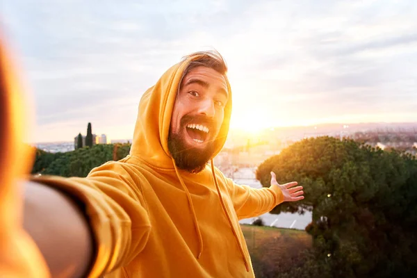 Selfie of young smiling man hooded sweatshirt. Happy Guy with good laugh taking self picture at sunset showing beautiful views. Cheerful traveler enjoying photographing their vacations having fun — Fotografia de Stock