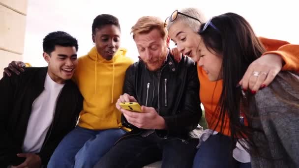 Group of Smiling people using mobile phone outdoors. Cheerful guys and girls friends having fun watching something at digital display. Happy vacations together. Social media and technology lifestyle — Vídeo de stock