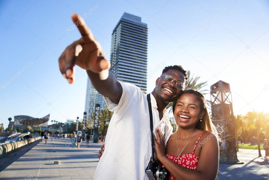 Smiling young African-American traveling couple enjoy a summer vacation in a resort town. The boyfriend points out something on the horizon to his pretty happy girlfriend . High quality photo
