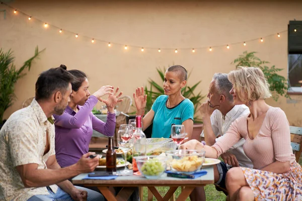 Group of friends chatting during lunch in the backyard. Middle-aged people having a discussion outdoors at the table while eating and drinking. Lifestyle and community concept. High quality photo