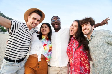 Portrait of five young multicultural friends on the sea shore looking at camera laughing. Multiracial young women and men strolling along a beach. High quality photo