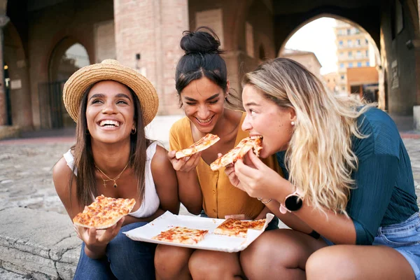 Three funny multiracial women eating pizza outdoors in the street. Happy female girlfriends enjoying the summer vacations together European city. Young people lifestyle concept. High quality photo