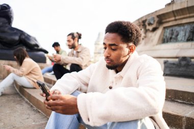 African American guy with mobile cell sitting in stairs of a city with other friends ignoring each other using smartphone. Technology addicts group of people. High quality photo