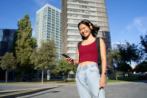 Young Happy Asian Woman Looking at Camera Using Phone. Smiling People with Headphones at city. High quality photo