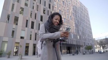 Business woman in a street urban corporate landscape using phone with electric scooter. Smiling healthy worker in eco transport. High quality 4k footage