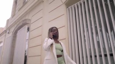 Cheerful African American business woman with braids walks through old city streets talking on cell phone via roaming connection. Happy excited young woman calling via smartphone. FullHD footage