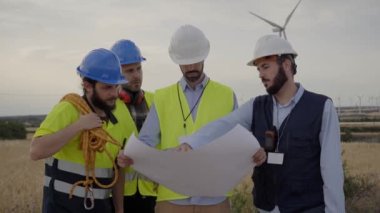 Sert şapkalı bir grup mühendis, rüzgar türbinleri olan bir tarımsal alanda plan üzerinde konuşup çalışıyor. Rüzgar santrali. Yenilenebilir ve temiz enerji kavramı, sürdürülebilir gelecek.