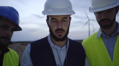 Bir grup miğferli mühendisin günbatımında yel değirmenlerinin arka planına karşı elektronik tablet kullanırken çekilen görüntü. Kavram: yenilenebilir enerji, teknoloji, elektrik, hizmet, yeşil, gelecek