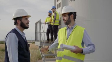 Sert şapkalı bir grup mühendis, rüzgar türbinleri olan bir tarımsal alanda plan üzerinde konuşup çalışıyor. Rüzgar santrali. Yenilenebilir ve temiz enerji kavramı, sürdürülebilir gelecek.