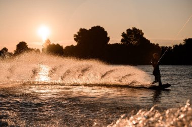 Aktif erkek sürücünün güzel manzarası halat tutuyor ve gün batımında su yüzeyinde sörf tahtasında..