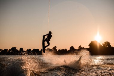 Aktif erkek sürücünün karanlık siluetinin harika görüntüsü halat tutuyor ve günbatımında wakeboard üzerinde aşırı atlıyor..
