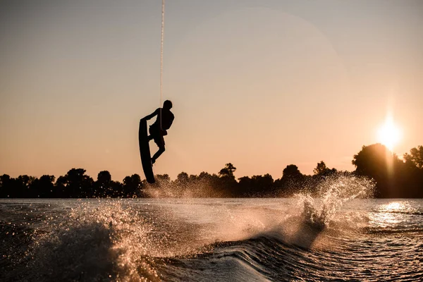 Vacker utsikt över mörk siluett av aktiva manliga ryttare håller rep och göra extrema hoppa på wakeboard vid solnedgången. — Stockfoto