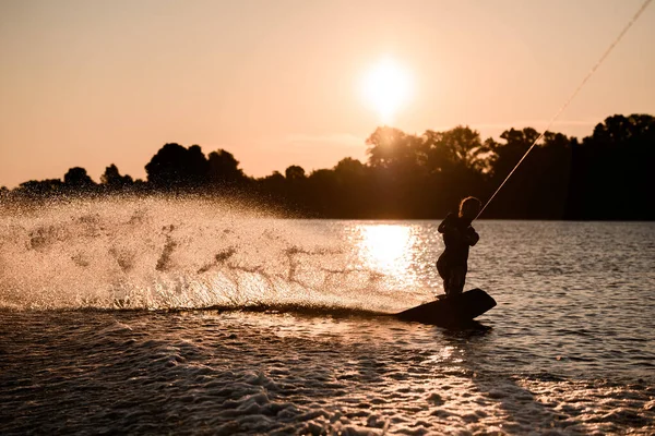 アクティブな男性ライダーの暗いシルエットの素晴らしい景色は日没時に水面にウェイクボード上のロープに乗って保持しています. — ストック写真