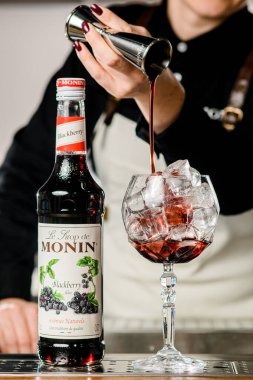 UKRAINE, KYIV - 12 Mart 2021: Bar tezgahında ve barmenin elinde bulunan Monin şurubu şişesi jigger 'dan buz küpleriyle şarap bardağına içilir
