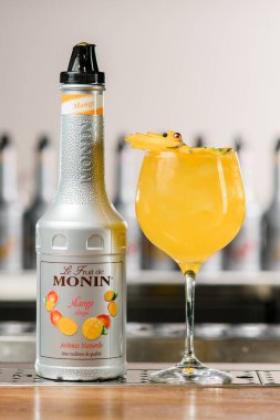 UKRAINE, KYIV - 12 Mart 2021: bir şişe Monin mango şurubu ve meyve dilimleriyle süslenmiş bir bardak parlak lezzetli kokteyl