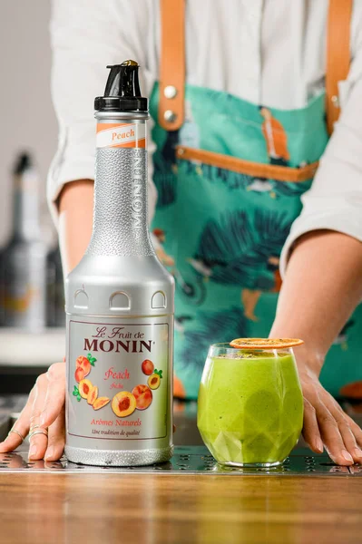 UKRAINE, KYIV - 12 maart 2021: vooraanzicht van fles Monin perzikstroop en glas groene cocktail met chiazaad en sinaasappelschijfje — Stockfoto