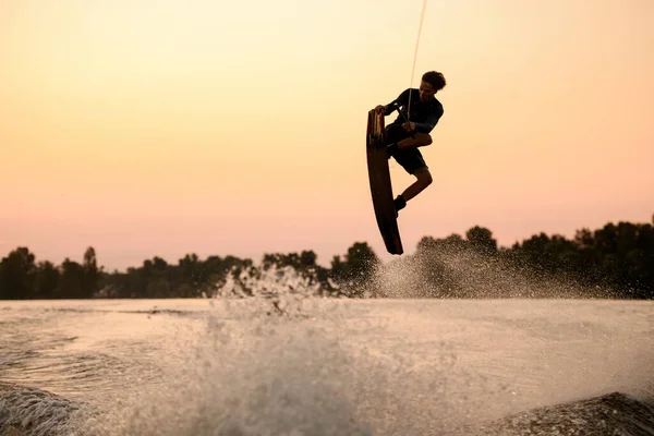 Silueta aktivního muže letícího přes stříkající vlnu na wakeboardu držícího se lana — Stock fotografie