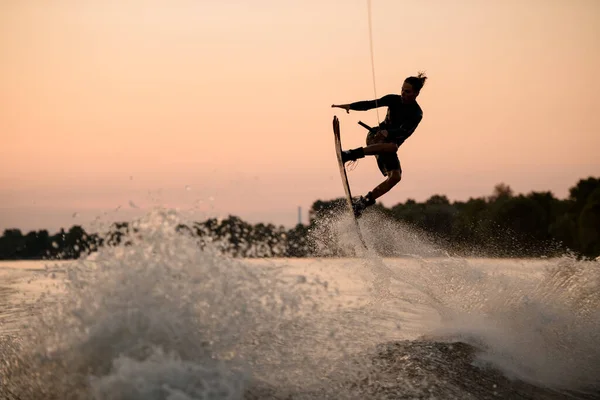 Ragazzo attivo che salta sopra l'onda spruzzante sul wakeboard aggrappandosi alla corda. Attività di sport acquatici. — Foto Stock