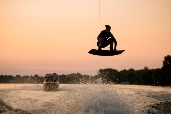 Uomo attivo che sorvola l'onda spruzzante sul wakeboard aggrappandosi alla corda. Attività di sport acquatici. — Foto Stock