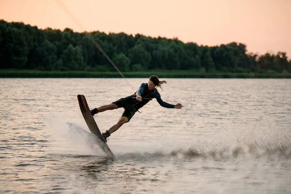 Schöner Kerl hält Seil und fährt energisch Wakeboard auf plätscherndem Flusswasser. — Stockfoto