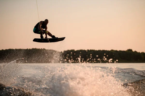 Homem muscular ativo pulando alto com wakeboard sobre a água salpicando contra o pano de fundo do céu — Fotografia de Stock