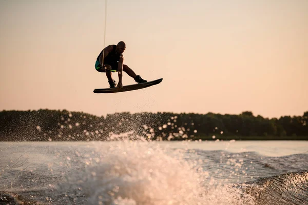 Muscular man jumping high with wakeboard over splashing water against the backdrop of sky — Stock Photo, Image