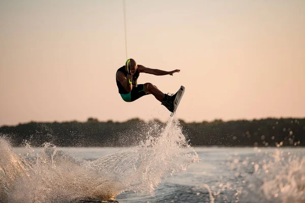 Healthy muscular man holding rope and jumping with wakeboard over splashing water — Stock Photo, Image