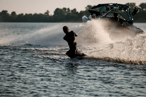 Atletische sportman houdt touw vast en balanceert op wakeboard achter motorboot op spetterende riviergolven. — Stockfoto