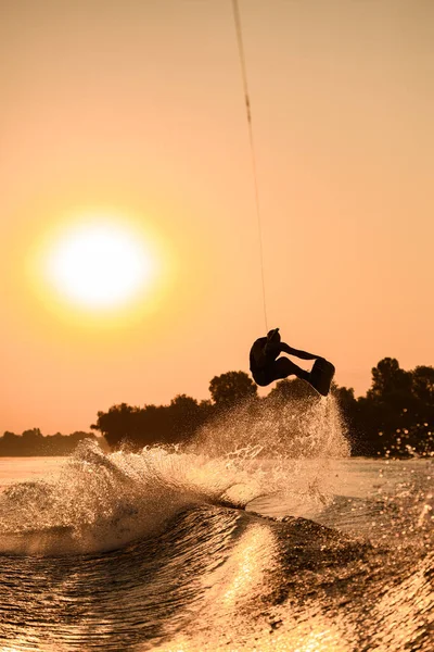 Homem ativo fazendo truque com wakeboard no céu laranja brilhante e fundo do sol. — Fotografia de Stock