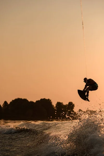 Excellente vue du cavalier masculin actif tient la corde et faire sauter sur wakeboard au ciel du soir sur fond. — Photo