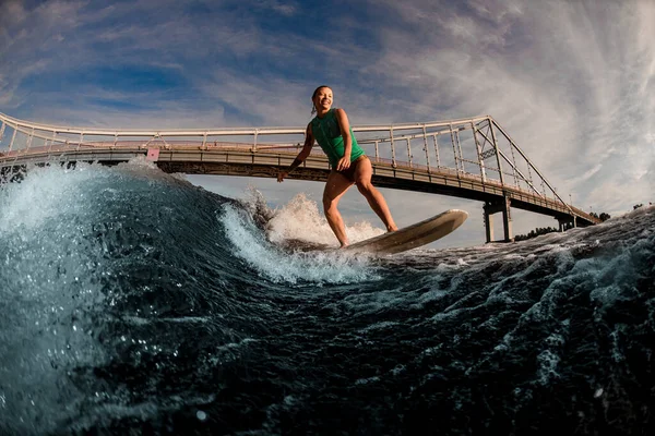 Leende kvinna wakeboarder skickligt rida ner våg på wakeboard mot bakgrund av bron — Stockfoto