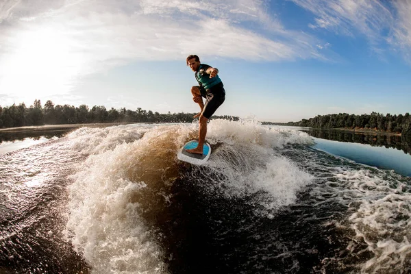athletic man makes stunts riding wakesurf on the blue splashing river wave against blue sky