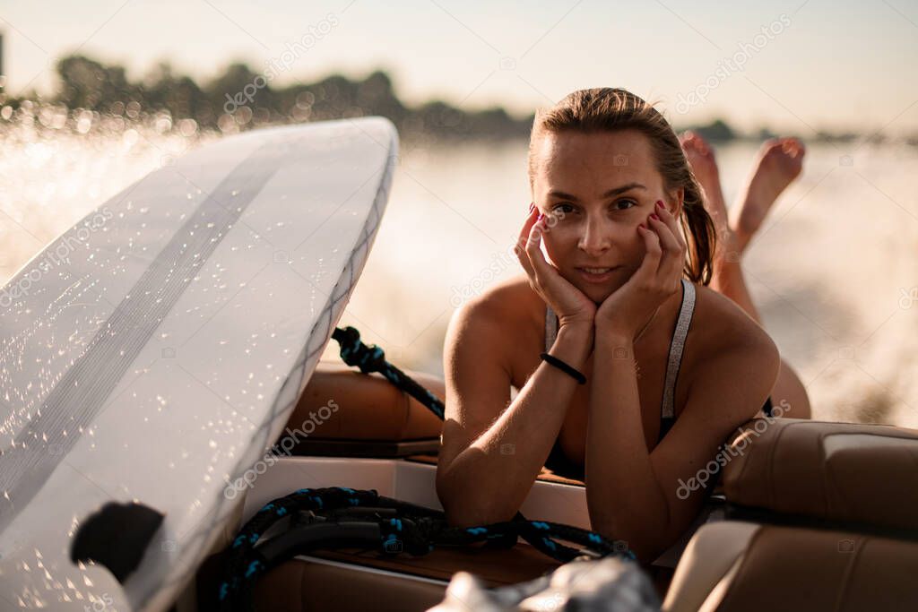 portrait of young woman in swimwear lying on edge of boat with wakesurf board