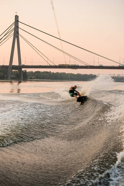 L'homme adulte fort tient la corde et équilibre énergiquement wakeboard sur éclaboussant l'eau de rivière — Photo