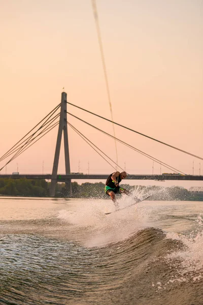 Homme actif tient la corde et saute habilement sur wakeboard sur fond de pont de la ville. — Photo