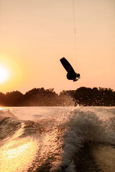 Ciemna sylwetka człowieka trzymającego linę i wykonującego skok na wakeboard o zachodzie słońca. — Zdjęcie stockowe