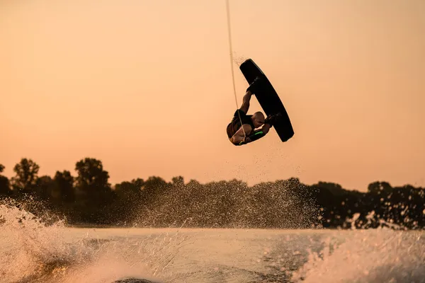Homme fort faisant astuce dans le temps de saut avec wakeboard sur fond de ciel coucher de soleil. — Photo