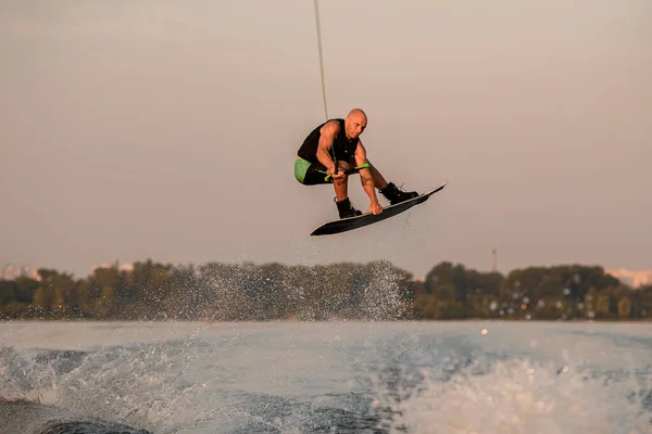 Uomo muscolare tiene cavo e saltando magistralmente sopra spruzzi d'onda sul wakeboard. — Foto Stock