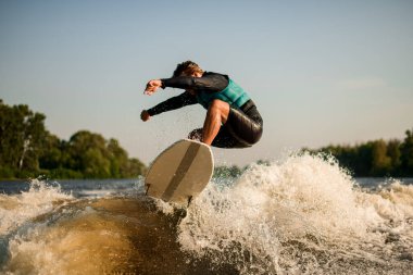 Esnek bir adam nehir dalgalarının üzerinde bir sörf tahtasına atlıyor.
