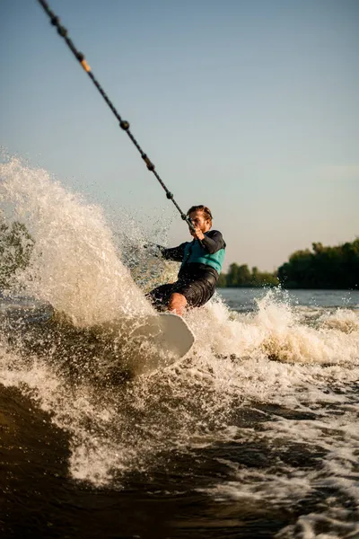 Sportlicher Typ hält Seil und reitet schnell auf Wakeboard auf plätschernder Welle. — Stockfoto