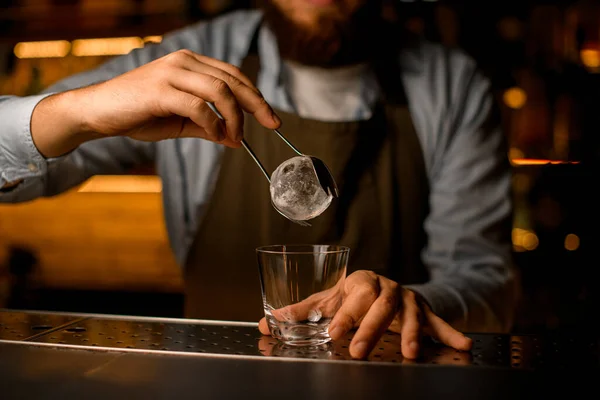 Mano del camarero sostiene pedazo redondo de hielo por pinzas sobre vidrio vacío — Foto de Stock