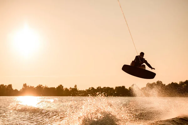 Grande vue de la silhouette du cavalier masculin tenant la corde et faisant un saut extrême sur wakeboard sur éclaboussures d'eau. — Photo