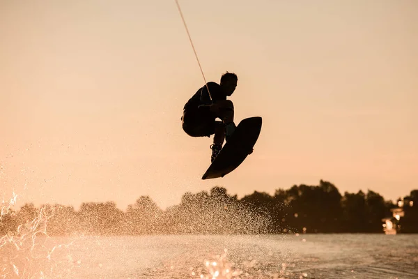 Silhueta escura do homem fazendo truque no tempo de salto com wakeboard no fundo do céu. — Fotografia de Stock