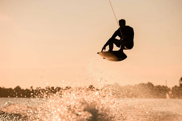 Vacker silhuett av manliga ryttare hålla rep och göra extrem hoppa på wakeboard över stänk vatten. — Stockfoto