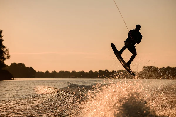 Silhueta maravilhosa de piloto masculino segurando corda e pulando no wakeboard sobre a água salpicando. — Fotografia de Stock