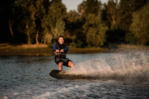Az atlétikai srác kötelet tart, és gyorsan lovagol a wakeboardon a fröccsenő vízfelszínen. Aktív és extrém sportok — Stock Fotó