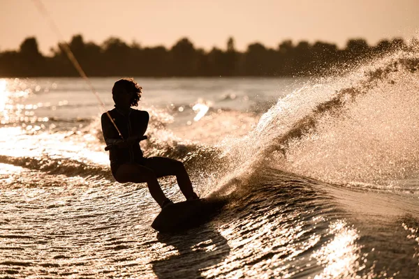 Silhouette of female rider holding rope and rides down on wakeboard on splashing wave. — Stock Photo, Image