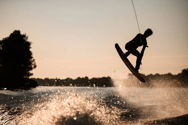 Silhueta de fêmea cavaleiro segurando corda e pulando alto no wakeboard sobre água salpicando. — Fotografia de Stock